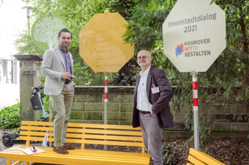 Zwei Personen an einer Bank. Einer hat ein Schild beschriftet.